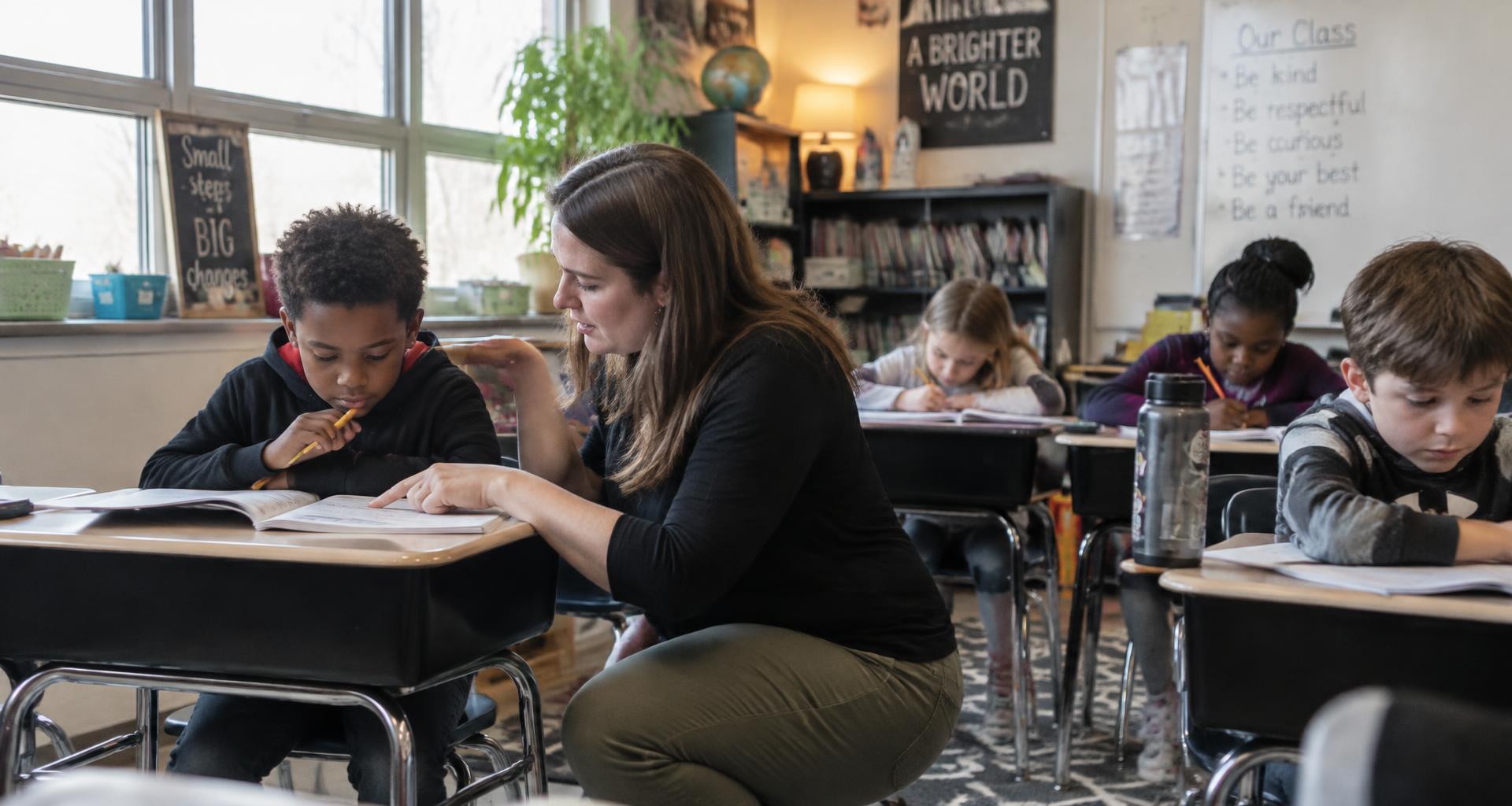 Teacher kneeling beside a student's desk giving individualized support while diverse classmates work independently